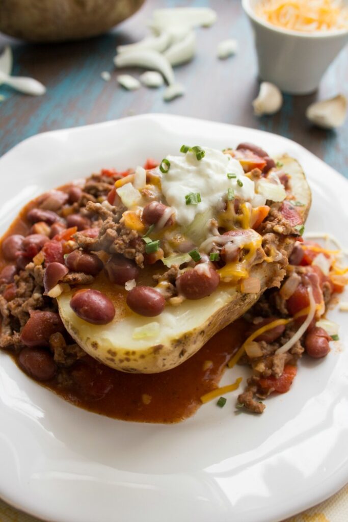 A loaded baked potato stuffed with chili and cheese ona white plate with more cheese and potatoes in the background