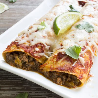Up close picture of a white plate containing 2 beef enchiladas topped with cheese, cilantro and a slice of lime