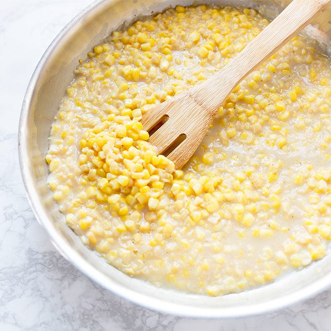 Wooden spoon stirring large stainless steel bowl of homemade cream corn