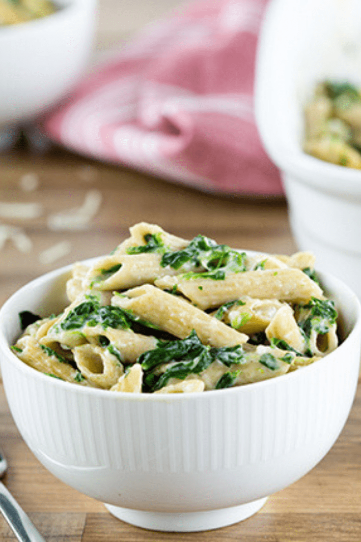A large white bowl of spinach pasta on a wooden surface with red cloth in the background