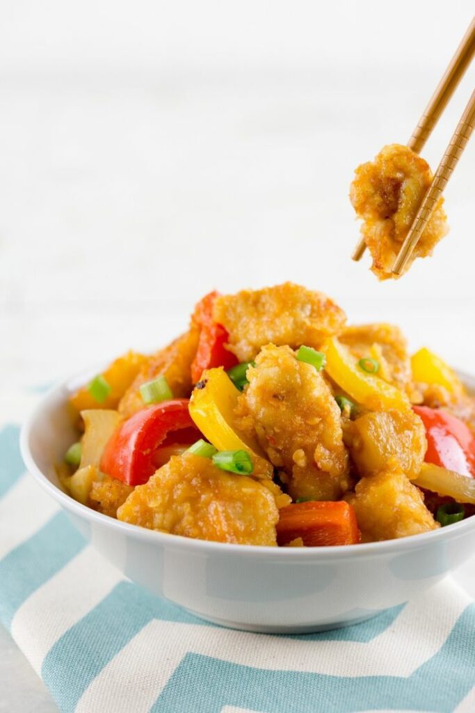 A white bowl of baked sweet and sour chicken with chopsticks holding a piece