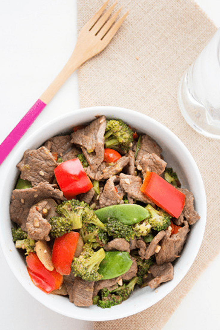 Overhead picture of a large white bowl of Chinese beef stir fry sitting on a burlap napkin with wooden knife in background